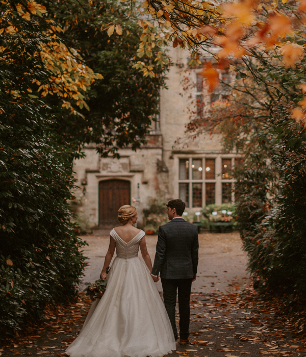 Bride and groom walking towards Minstead Lodge with autumn leaves in the distance