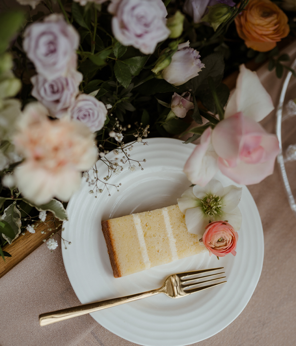 A slice of wedding cake on a plate, with a fork and surrounded by colourful flowers