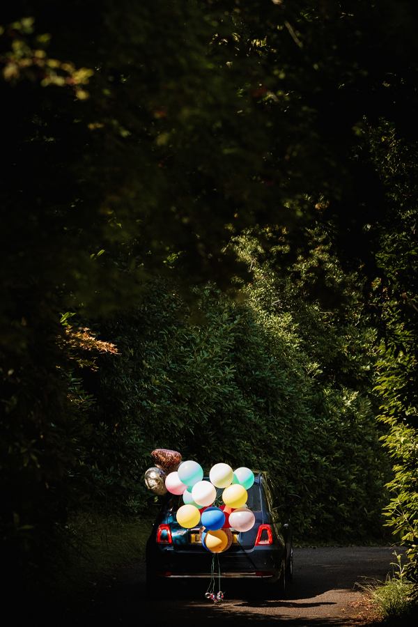 A blue car decorated with balloons driving under trees