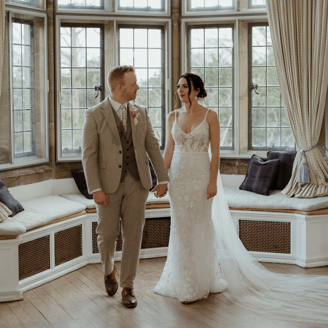 A wedding couple walking towards the camera in a bright room, holding hands.